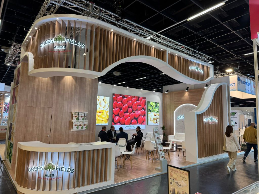 Modern exhibition stand for Green Fields Alex at Anuga 2025 featuring a curved wooden slat design and digital screens displaying fresh produce.